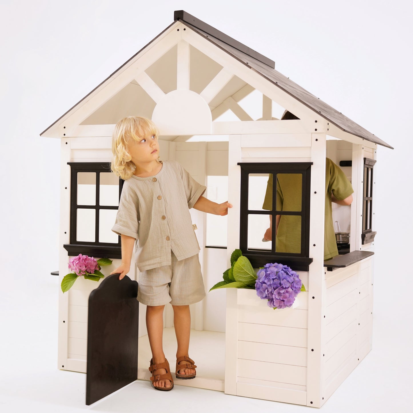 Blonde child standing in the doorway of a white wooden playhouse with black trim, holding the black gate-style door open.