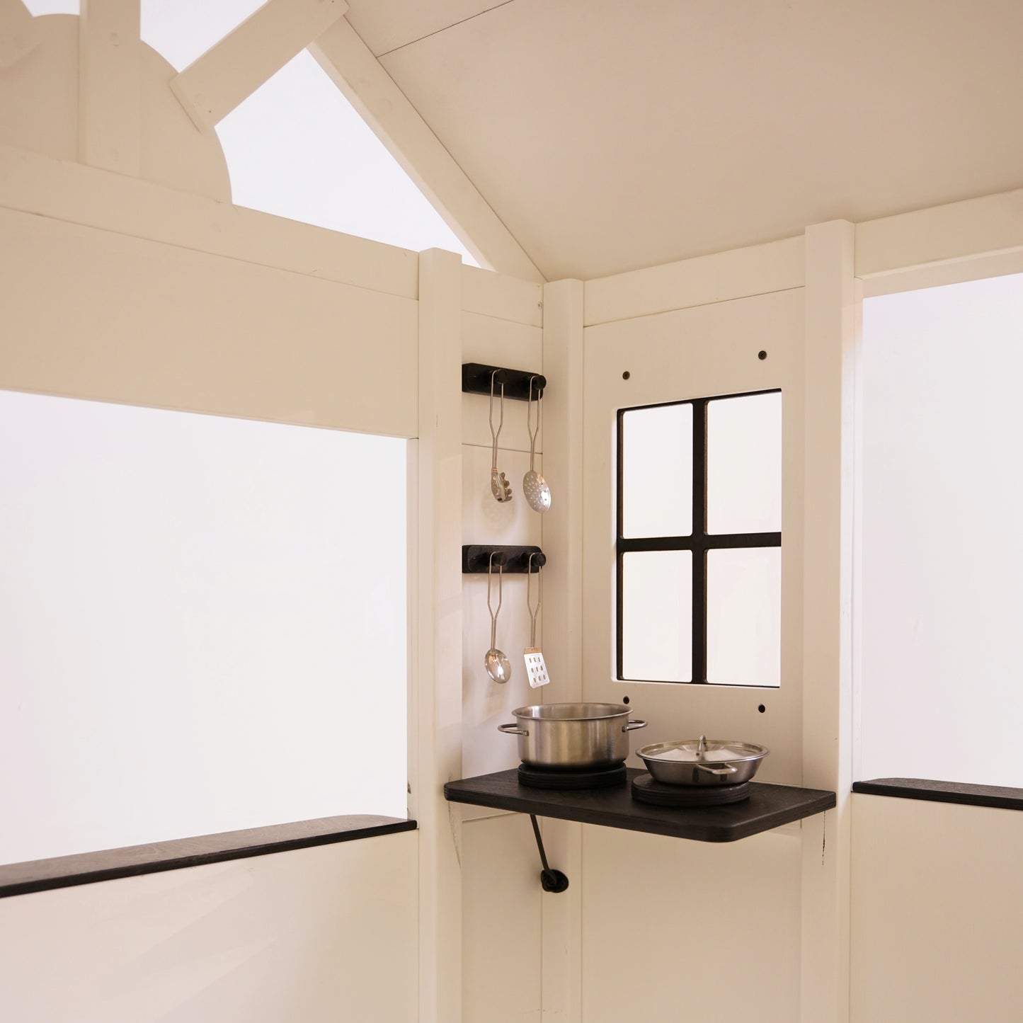 Inside view of a white wooden playhouse showing a black corner shelf, hanging utensils, and toy cooking pots.