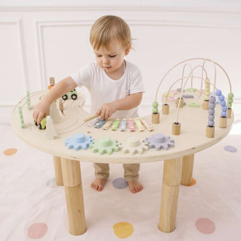 Child playing with a wooden activity table filled with colorful toys on a white floor.