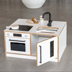 Children's play kitchen set with white surface, black stove, oven, and sink on a gray floor.