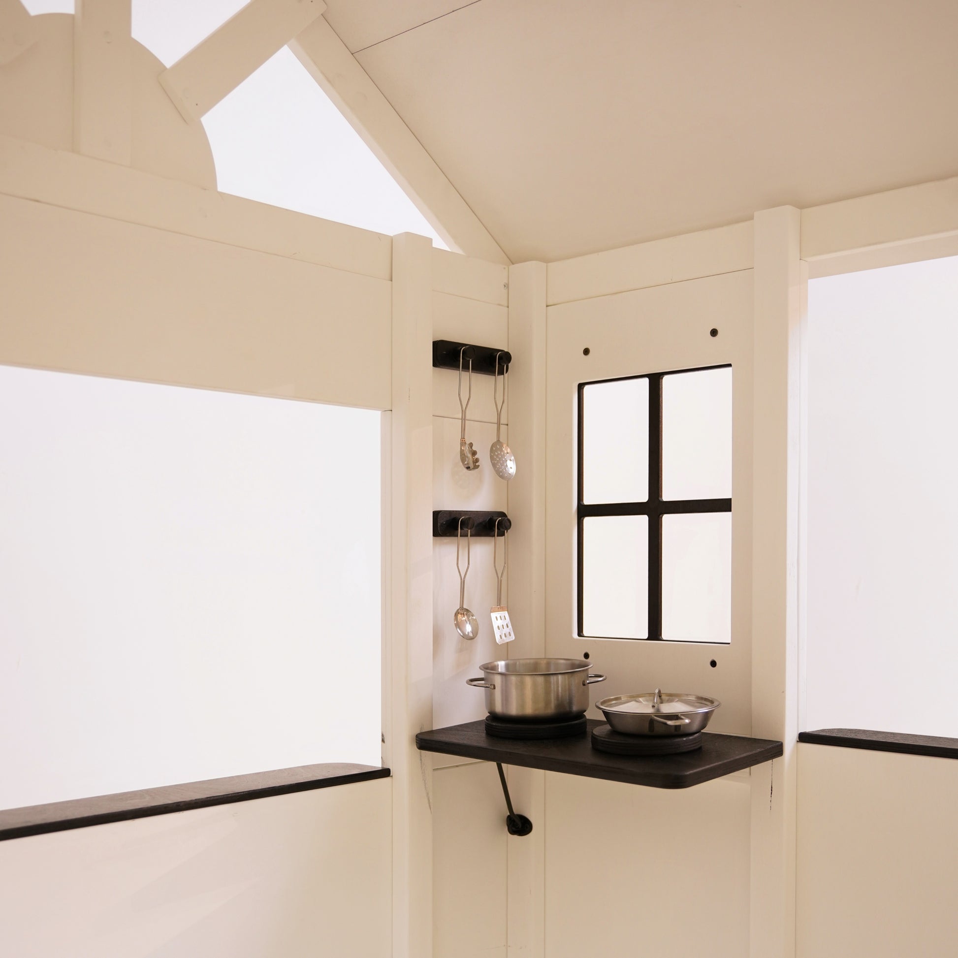 Inside view of a white wooden playhouse showing a black corner shelf, hanging utensils, and toy cooking pots.