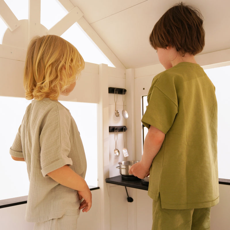 Two children playing inside a white wooden playhouse, standing at a small black countertop with hanging kitchen utensils.