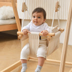Baby sitting in the Avenlur off white cotton canvas hammock swing seat with wooden front bar, hanging from a wooden indoor swing stand